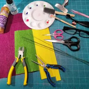 A desk from above. On top of the desk, it has various art supplies like crepe paper, paints, glue, scissors, pliers and brushes.