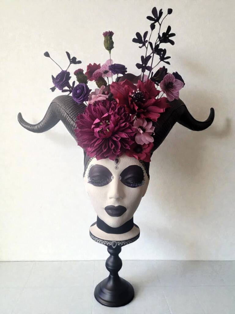 A Styrofoam head on a black pedestal with horns and a flower crown. Flowers include dahlia, ranunculus, Mochara orchids, and lisianthus. The head has very dark makeup around her eyes and lips.