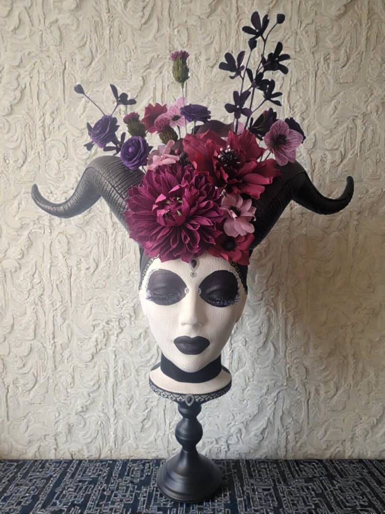 A Styrofoam head on a black pedestal with horns and a flower crown. Flowers include dahlia, ranunculus, Mochara orchids, and lisianthus. The head has very dark makeup around her eyes and lips.