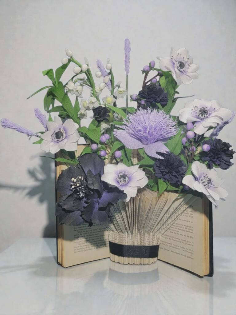 A book turned into a vase holding purple, white and black flowers.