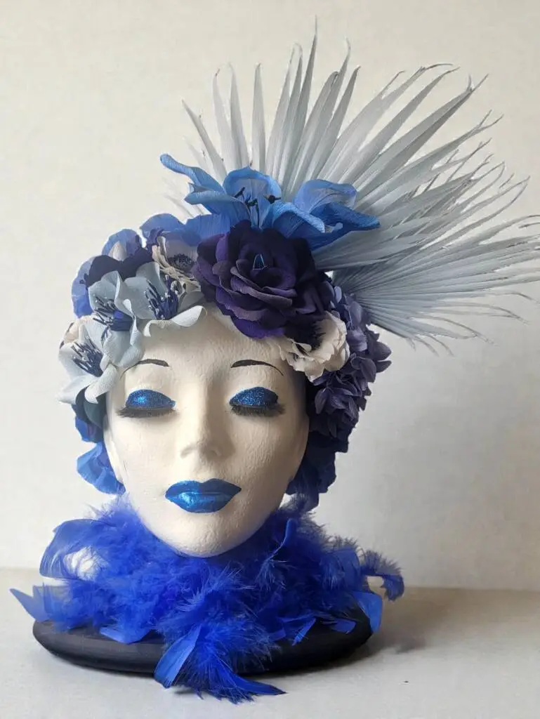 A Styrofoam form of a head with blue flowers as hair, blue glitter eye shadow and lipstick and a blue feather boa.