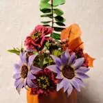 An orange pumpkin filled with purple sunflowers, orange calla lillys and greenery